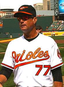 A man wearing a white baseball jersey with "Orioles" in orange script across the chest and the block number "77" in orange below it; he is also wearing a black baseball cap with an orange and black bird on the front and an orange brim