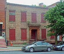 A two-story brick facade with red boards in the windows and brownstone around the entrance. Parked in front of a tree on the street is a dark gray late-2000s Honda Civic