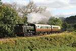 A green pannier tank locomotive is travelling from right to left, bunker first, along a single track on a grassy embankment. The locomotive is pulling two passenger carriages painted cream and brown, and a plume of smoke is passing over the first carriage. In the background are a number of green trees, but behind them are some taller trees that have lost most of their leaves.