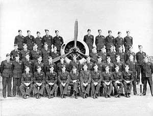 Four rows of men in dark military uniforms surrounding an aircraft's three-bladed propeller