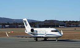 A twin-tailjet, high-tailplane passenger aircraft painted white above and grey below in front of an aircraft hanger and other buildings. A similar aircraft is visible in the background.