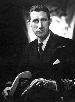 Black and white studio portrait of a sitting man in naval uniform. He has well groomed hair, two medal ribbons on his left breast, and is holding a cap in one hand by his knees.