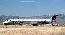 Right side view of an airplane taxiing on the ground towards left side of image. Another plane is behind it, and in the background are mountains and blue sky with a few clouds.