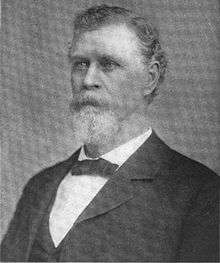 A man with gray hair, beard, and mustache wearing a black jacket and neck tie and a white shirt