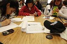 Three high-school age students sit around a round table. Two are writing letters on white paper and the third (on the right) is holding a water bottle. A packet of paper that is titled STAND sits on the middle of the table.