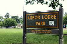 Wooden sign with text identifying the name, management, and hours of operation for the park; in the background is a green lawn, part of a building, and two people.