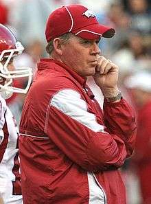 Coach Petrino in a red Arkansas jacket and hat on the field with a player at rear.