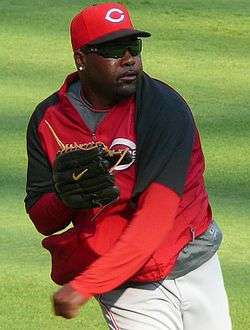 A dark-skinned man with sunglasses and a goatee follows through after throwing a baseball; he is wearing a red nylon warmup jacket and red baseball cap.