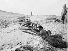 Black and white photo of a group of men wearing military uniforms crouched in a trench in rocky terrain. The men are all aiming rifles forward. Several men are standing behind the men in the trench.