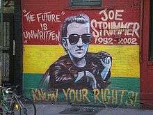 A photograph of the painting of the memorial mural of Joe Strummer on the wall of the Niagara Bar in the East Village in New York City. The mural depicts Strummer (centre) surrounded by the words "THE FUTURE IS UNWRITTEN" (on the left), "JOE STRUMMER 1952–2002" (on the right), and "KNOW YOUR RIGHTS!" (bottom) on a horizontal tricolour of red, yellow, and green background