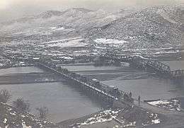 Photo shows a line of 1918-style trucks crossing a bridge. An nearby bridge is demolished.