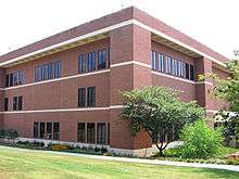 A three-story rectangular brick building seen from its corner. There is a small tree near its corner, and grass in the foreground.