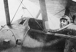 Close-up photograph of the side of an aeroplane. A man wearing a cap and with goggles on his forehead is seated in the cockpit.