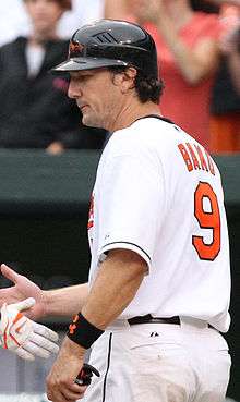 A man in a white baseball uniform faces to the left. He reaches to shake hands with another unseen person. His uniform reads "Bako" in small orange print and "9" in larger orange print on the back, and he is wearing a black batting helmet with an orange-and-black bird on the face.