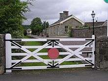 An ornate white gate obscures the view of a small stone station building and platform, with grass growing where the railway tracks would have been. In the distance at the far end of the platform is the original signal building.