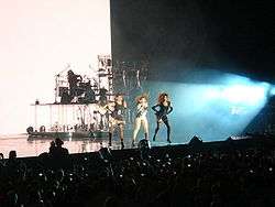 A long shot of three women performing in front of a crowd. From left to right, the first and third are dancing, while the second is singing. Behind them, a band, playing guitars and drums, is visible. The women are wearing black dresses.