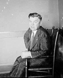 A black-and-white photo of a man with thick, dark, combed-over hair seated in a wooden chair with a pained look on his face