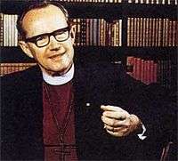 "Bo Giertz, a middle-aged man with dark-framed glasses, is seen in his office in front of a wall of books on shelves; he is wearing a plain white clerical collar, maroon shirt, large cross and black suit. His left arm is in the foreground; he is animated as if trying to make a point."