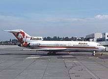 Right side view of an aircraft parked on the tarmac, with a tree-covered hill as well as some clouds and buildings in the background