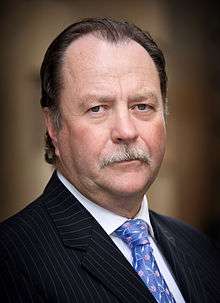 A Caucasian male in his late fifties wearing black pinstripe suit, white shirt and blue decorated tie.