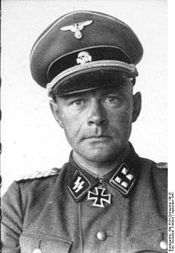 A man wearing a military uniform, peaked cap and a neck order in the shape of a cross. His cap has an emblem in shape of a human skull and crossed bones.