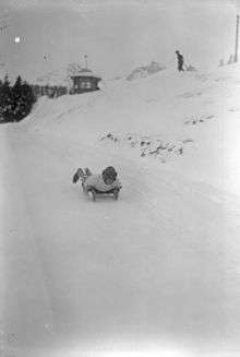 An athlete prone on a sled, going down a course. A small building, trees, and another person can be seen in the near background and snowy peaks in the distance.