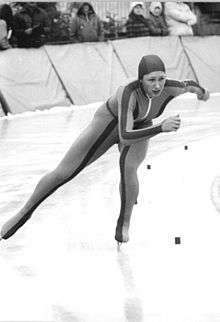 A woman is wearing a skin-tight jumpsuit with a hood and ice skates; she is making a turn as she skates on an ice track.