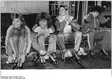 Four women wearing tracksuits and speed skates are sitting on a bench. Three of them are smiling, and among these, two are tying up their skates while a third one is clapping her hands.