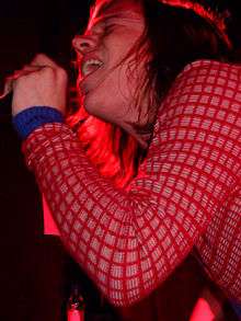 A long-haired Caucasian man sings into a microphone, illuminated in a red light.