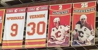 Four rectangular banners are hanging from the rafters in a hockey arena. The two on the far left are predominantly white with red and gold trim. The left one of these says "1981–1989 McDONALD 9" and the right "1982–2002 VERNON 30". On the right side of them, two more banners are shown. Both are mainly red, each showing (from top to bottom) a number, then a red banner with gold trim showing a name, and lastly a person in full hockey gear, who is shown wearing a white jersey with red trim and a white helmet. The left one of these says "2 MacINNIS", while the right one says "25 NIEUWENDYK".