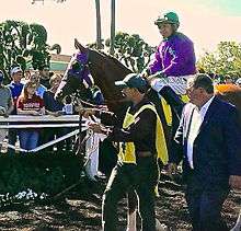 A racehorse and jockey being led by a groom, with a man in a business suit walking alongside