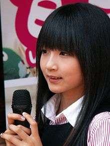 A black-haired girl sings while holding a microphone with both hands.