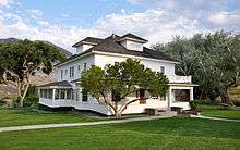 A white two-story house with front and side porches and a widow's walk rests on a property with green lawn, trees, and a picnic table.