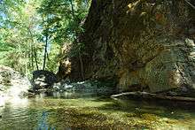 A pool along a steep, gorge-like portion of the Carmel River is illuminated by the sun with layers of colors showing themselves through the water.