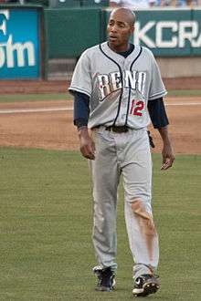 A dark-skinned young man wearing a gray baseball uniform walking across a grass field