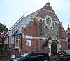 Three-quarter view of a steep-roofed brick building with large areas of stone dressing, especially on the entrance porch. The façade has two tall and two shorter pointed-arched windows and a round window, all with green tracery.  The side wall has large rectangular windows.