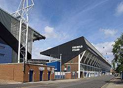 A large association football stand with part of a floodlight on the left-hand side and road and tress on the right-hand side