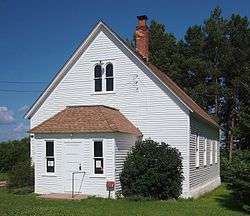 Summer view of simple wooden building with gable roof and projecting entrance vestibule