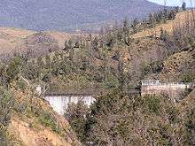 Waterfall surrounded by stone walls in brown wooded hills; many of the standing trees are barren, gray, and dead.