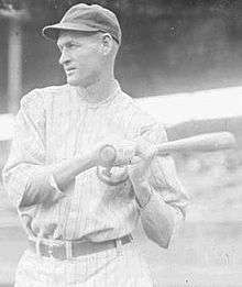 A black-and-white photograph of a man wearing a white pinstriped baseball uniform and holding a baseball bat over his shoulder with both hands