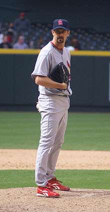 A man with a dark goatee standing on a pitcher's mound, wearing a gray baseball uniform with red trim and a navy-blue baseball cap