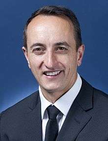 Official photograph of middle aged man with combed-back short black hair and no other facial hair, wearing a suit, smiling into the camera.