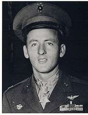 Head and shoulders of a young white man with a peaked cap pushed high up on his forehead, wearing a military jacket with two rows of ribbon bars on the left breast and a star-shaped medal hanging from a ribbon around his neck.