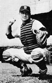 A crouching man wearing a catcher's chestguard and baseball cap prepares to throw a baseball with his right hand