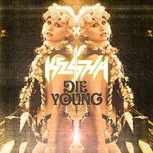 A Caucasian female with blond braids and a floral diadem. She is sparsely clothed in a suit of leather strips with much skin expose. Back facing a mirror, her reflection is symmetrically depicted with the words "Kesha" and "Die Young" appearing in the center.