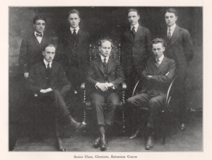 Seven men, three seated and four standing behind the three, look straight ahead with solemn expressions. Formally dressed in suits and ties, the men are about age 20.