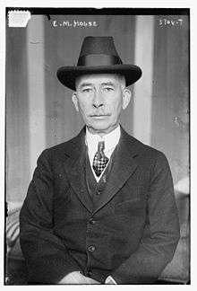 Frontal image of man with white mustache, white shirt, necktie, dark suit and hat. He appears to be seated with hands folded in his lap.