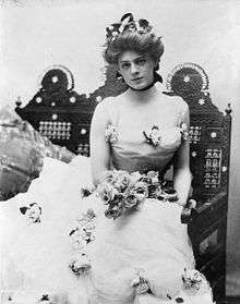 Ethel Barrymore, as a beautiful young woman, in a three-quarter length portrait, seated, facing front, wearing an elaborate gown and holding a bouquet of roses