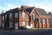 A dark red brick hall on a corner site, with adjoining houses in an identical style. The hall, partly obscured by a traffic light, has a pointed-arched entrance with a wooden door below a rectangular window frame with three lancets.  To the right of the door, there are three plain windows between brick buttresses.  Above the entrance is a steep roof perpendicular to the main roof.