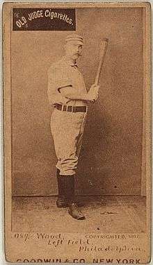 A sepia-toned baseball card image of a man wearing an old-style white baseball uniform and pillbox cap holding a baseball bat with both hands